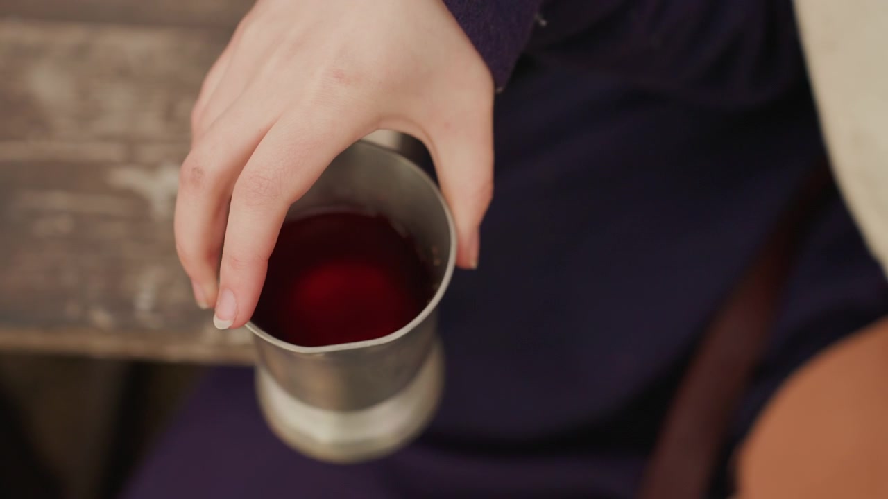 Primo piano dall'alto di una mano adulta che inclina una tazza di metallo piena di liquido rosso scuro, mostrando un'azione di versamento attenta vicino al bordo, caldi riflessi metallici e luce soffusa su una scena intima da tavolo.
