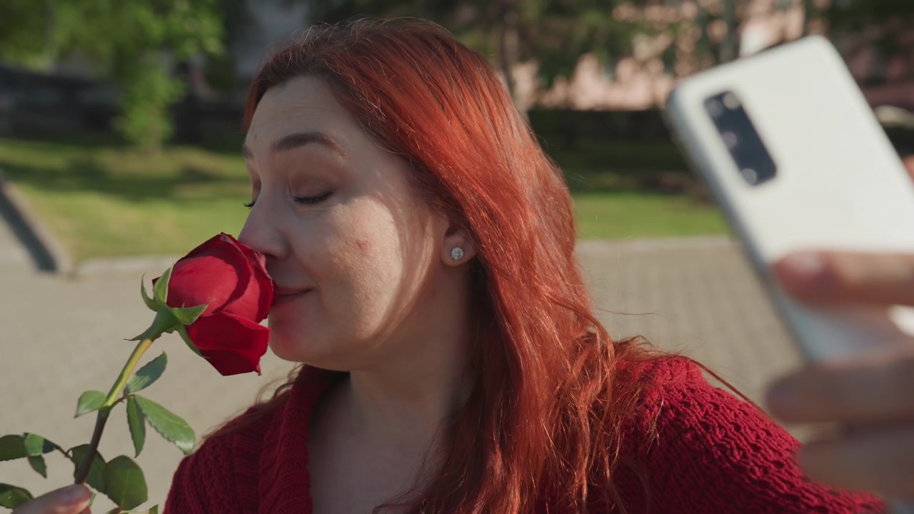 Primo piano di una donna con lunghi capelli rossi che indossa un maglione a maglia testurizzato, mentre tiene e annusa una vivace rosa rossa e si scatta selfie all'aperto su un terreno pavimentato con alberi verdi in luce soffusa.