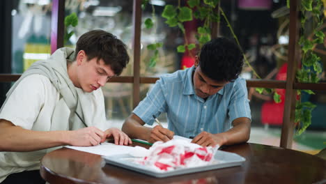 two colleagues bend over desk intensely drawing varied sketches on paper while hands guide pencils and pens in focused creative session within bright modern office studio environment atmosphere vibe