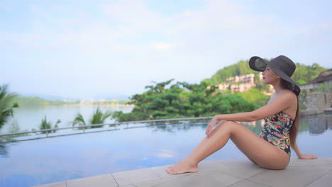 Mujer-Asiática-Solitaria-En-Traje-De-Baño-Y-Sombrero-Flexible-Sentado-Junto-A-La-Piscina-Disfrutando-De-La-Vista-Del-Paisaje-Tropical,-Cámara-Lenta-De-Marco-Completo