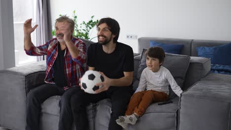 Three-generations-of-men-relax-on-couch-in-living-room-watch-football-match