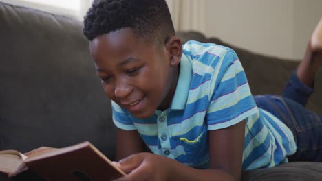 Niño-Afroamericano-Leyendo-Un-Libro-Mientras-Está-Acostado-En-El-Sofá-En-Casa