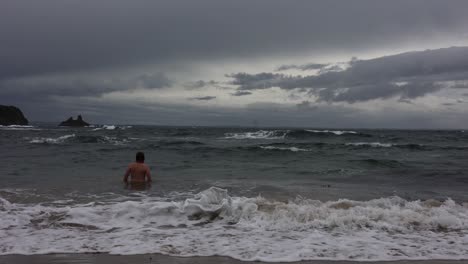 a slow motion shot of a beareded man walking into stormy waves and swimming in winter