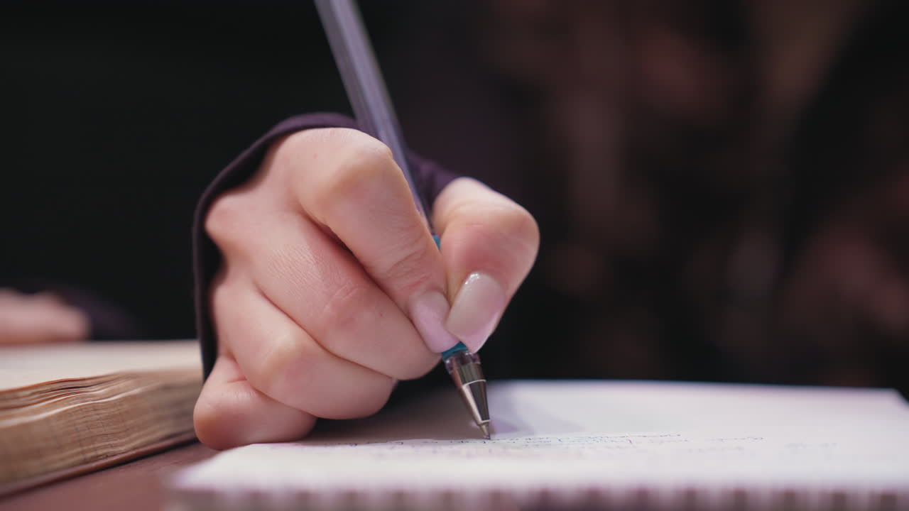Close up of female hand writing with blue pen featuring silver tip on spiral notebook. Fingers gracefully grip pen, smooth acrylic nails visible, warm tone background emphasizes quiet focus and detail