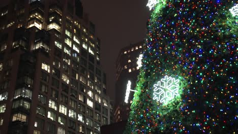 Close-up-parallax-of-Vancouver-city-very-large-and-tall-christmas-tree-lit-up-and-glowing-surrounded-by-the-tall-lit-up-buildings-and-skyscrapers-at-on-a-winter-night-in-the-dark