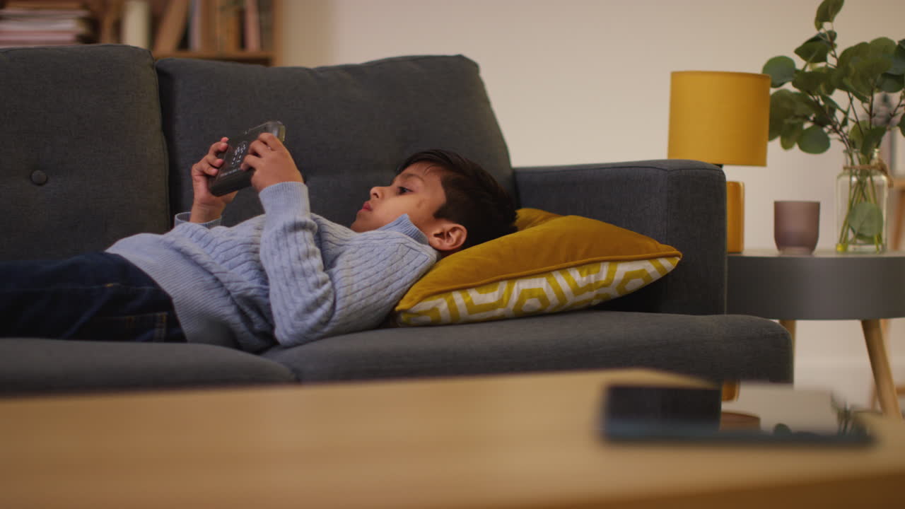 Young Boy Lying On Sofa At Home Playing Game Or Streaming Onto Handheld Gaming Device 7