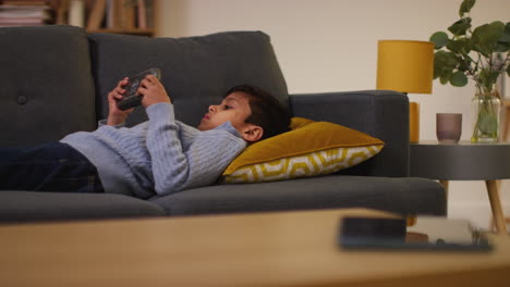 young boy lying on sofa at home playing game or streaming onto handheld gaming device 7