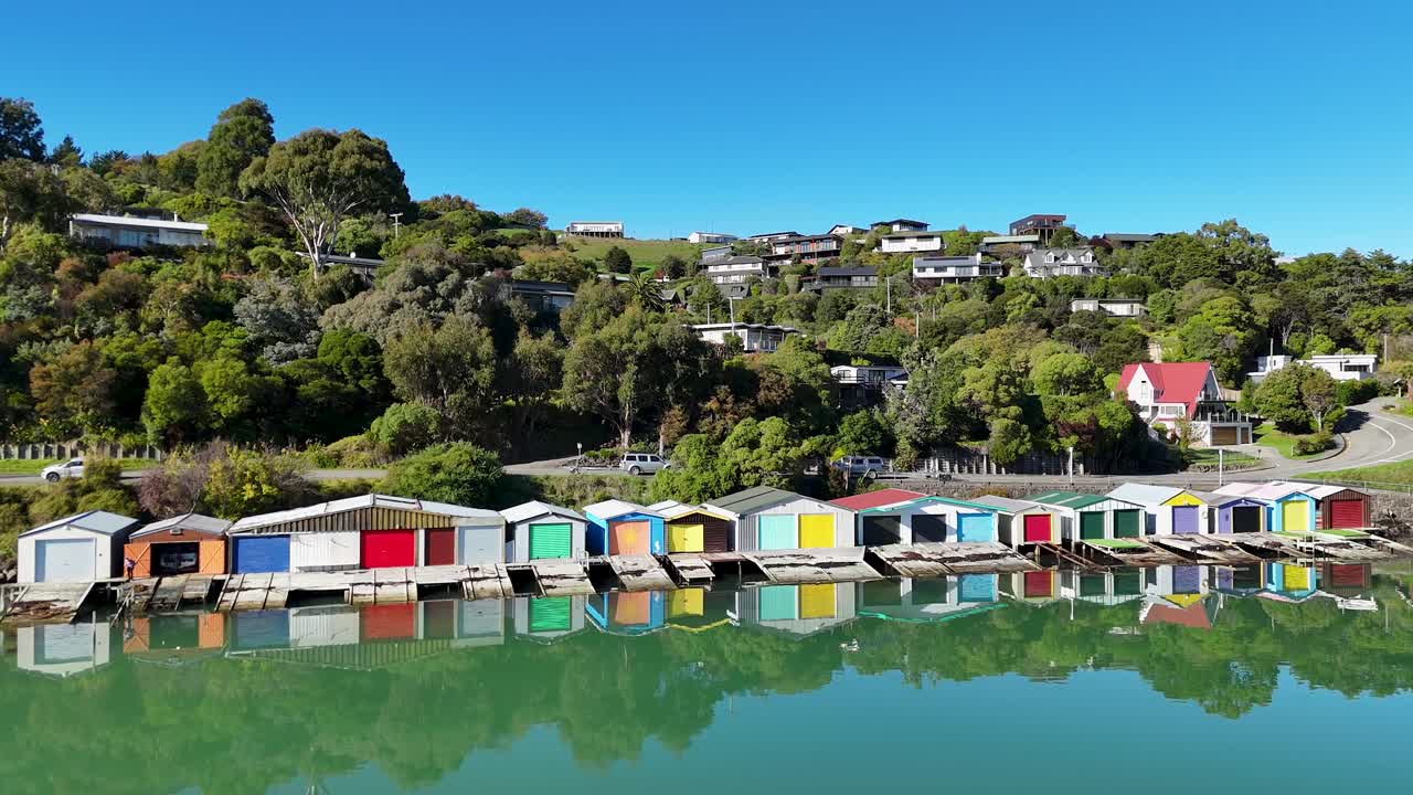 Vibrant boat houses line the serene waters of Akaroa, reflecting vivid colors under clear blue skies