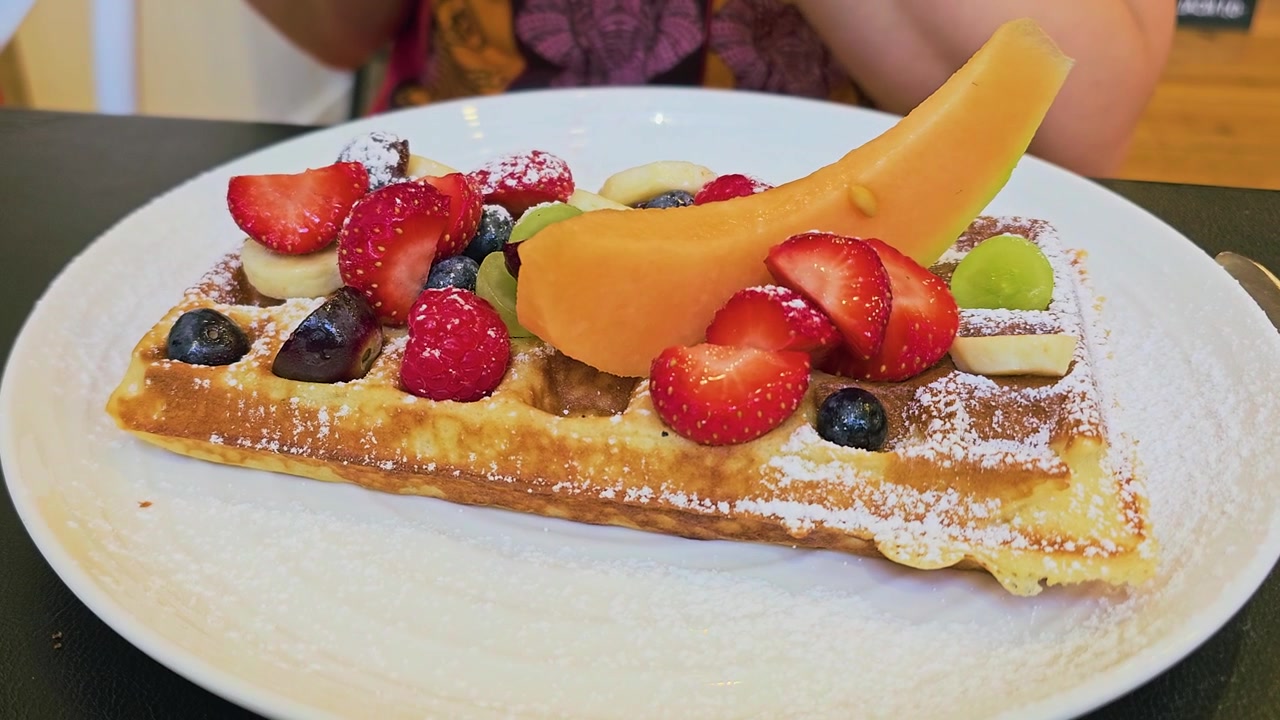 Waffle ricoperto di frutta fresca con zucchero a velo, fragole, lamponi, mirtilli, banana, uva e melone, servito come colazione dolce o dessert all'interno di un accogliente caffè