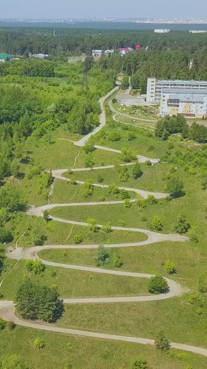 hairpin curves of path on beautiful green hill with forest against buildings on sunny autumn day aerial view