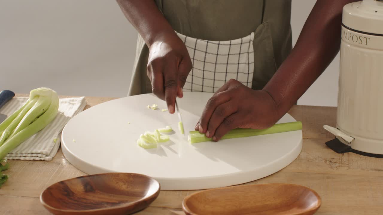 Mid-adult man slicing celery at kitchen counter on white board with chef's knife, wearing apron