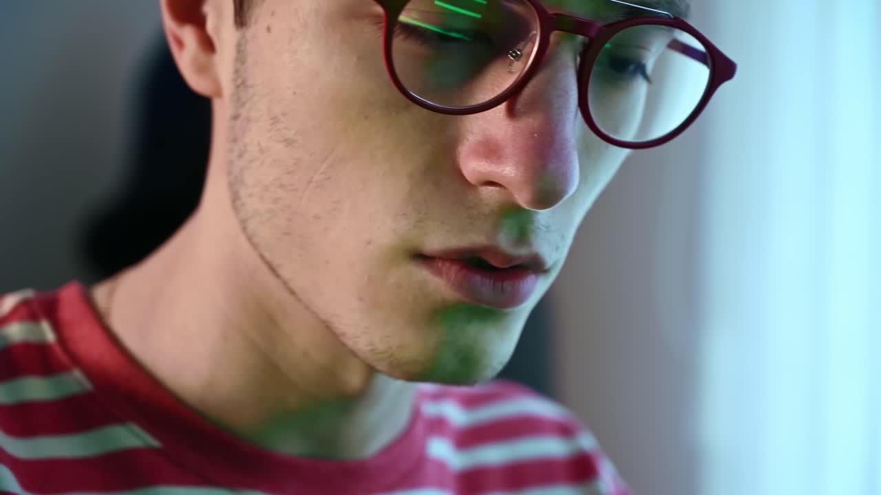 Young pimply man in red glasses and pink-striped sweater writing codes on a green page in his computer. Concentrated programmer. Reflection in the glasses. Side view. Close up