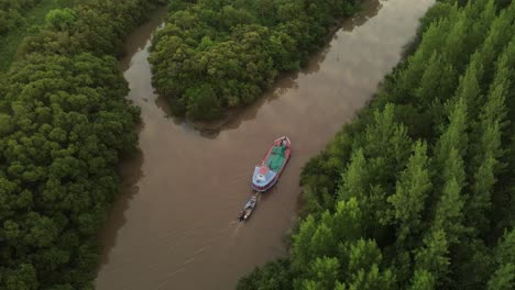 cinematic drone orbit shot of ship carrying small boat on amazon river surrounded by green rainforest trees during sunset