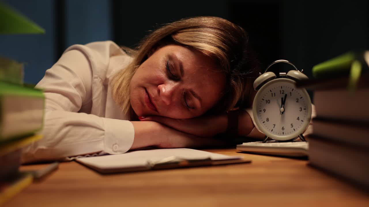 Tired woman sleeping on a desk amidst books and an alarm clock