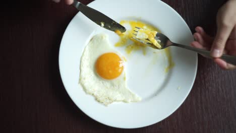 Top-view-of-hand-with-knife-and-fork-eating-fried-egg-on-plate