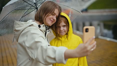una feliz mujer rubia con una chaqueta blanca toma una selfie con su hija adolescente en una chaqueta amarilla que hace caras y posa con un paraguas después de la lluvia en el parque durante un paseo
