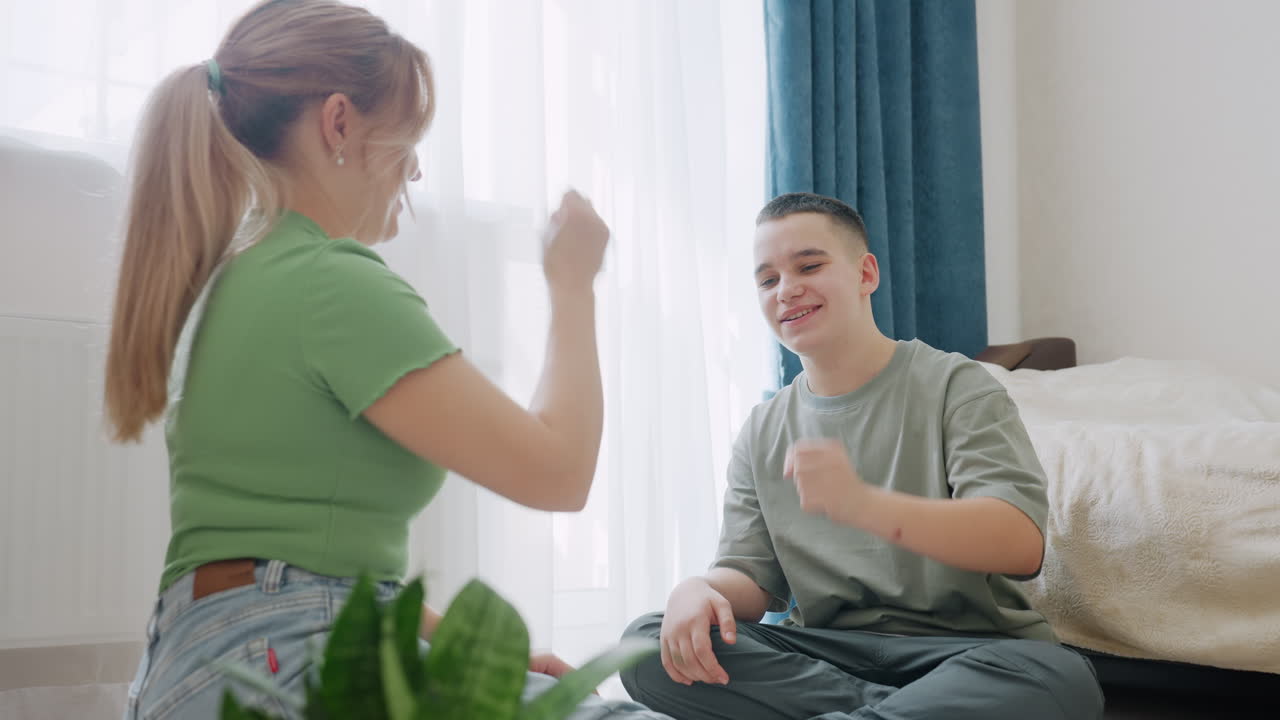 Smiling mother and son sitting on floor playing cheerful hand game in bright cozy room filled with natural sunlight, enjoying relaxed bonding time, laughter, and emotional connection
