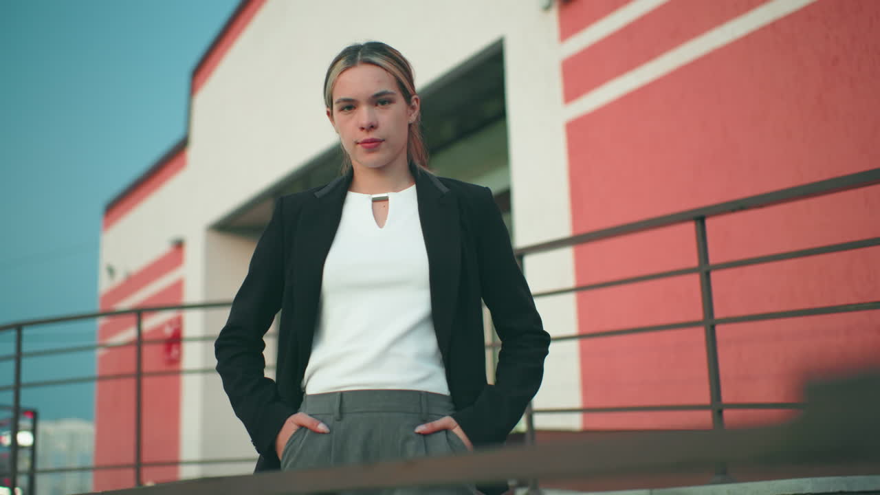 Confident woman in black blazer and white top poses with hands in pockets near iron railing in front of modern office building under clear sky, radiating authority, and professionalism outdoors