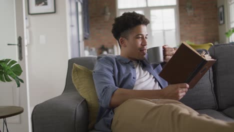 relaxed biracial man sitting on sofa in living room reading book and drinking coffee