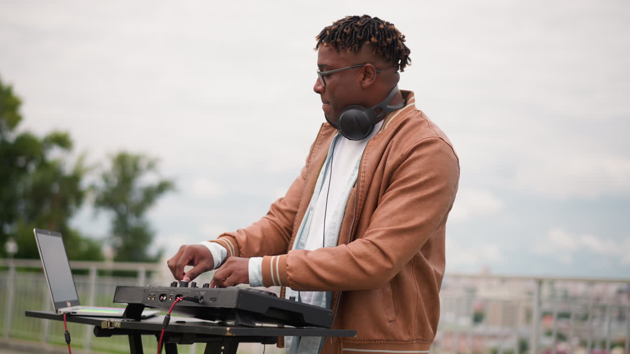 black DJ playing rooftop street set, closeup profile and fullbody shots, headphones around neck, hands on mixer and turntable, laptop open on stand, city skyline and cloudy sky in background