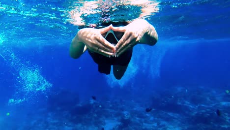 mujer caucásica buceando en un mar azul claro entre peces mientras hace un símbolo de amor con sus manos