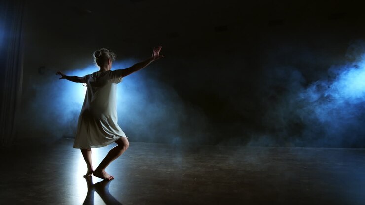 Uma mulher de vestido branco dança no palco as danças dramáticas do balé moderno. Uma bailarina realiza movimentos plásticos, executando coreografia moderna em meio à fumaça.