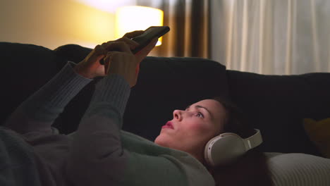 woman wearing wireless headphones lying on sofa at home at night streaming or looking at online content on mobile phone 2