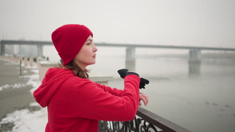 woman in red hoodie and black leggings rests hands on decorative metal railing by calm river during winter, snow-covered ground, distant bridge with moving vehicles, misty atmosphere