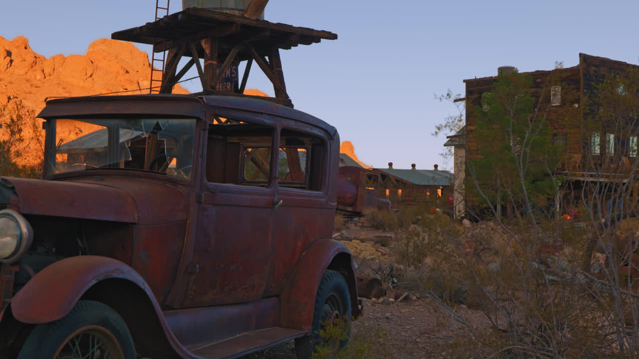 An old rusty truck sits in a desert ghost town amongst old buildings and a desert landscape.
