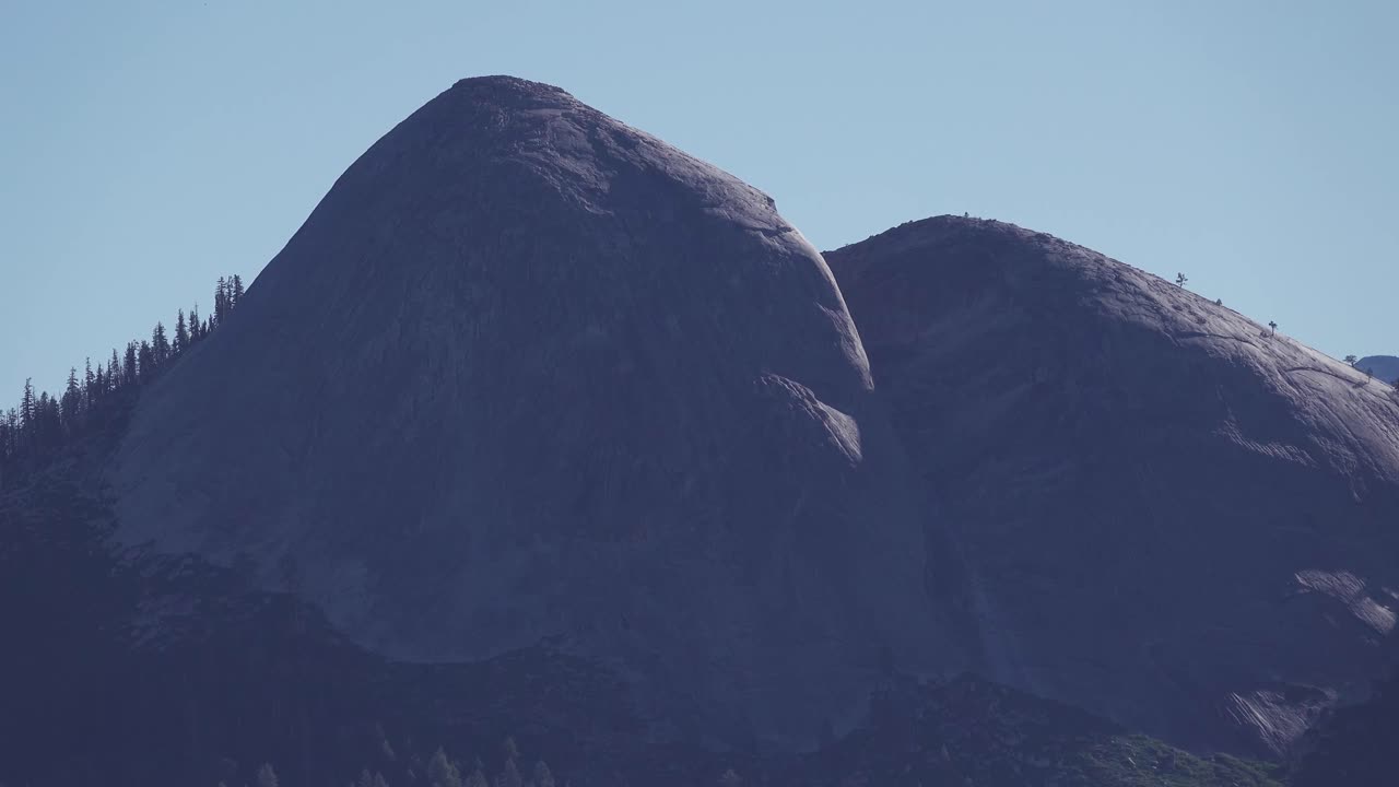 Lenta ripresa con zoom indietro di enormi cupole di granito che si ergono dal paesaggio montano nell'alta Sierra, rivelando aspre formazioni naturali sotto cieli limpidi