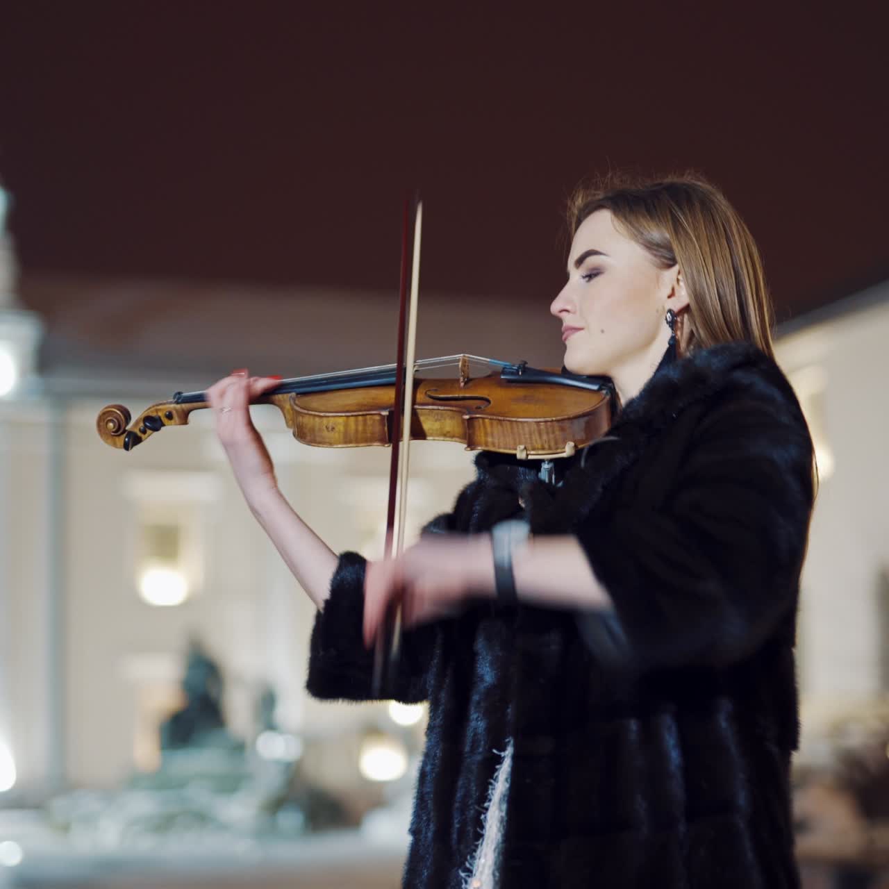 A romantic girl in warm clothes is playing a violin in the street in the square on the background of urban architecture in the evening. Blurred background.