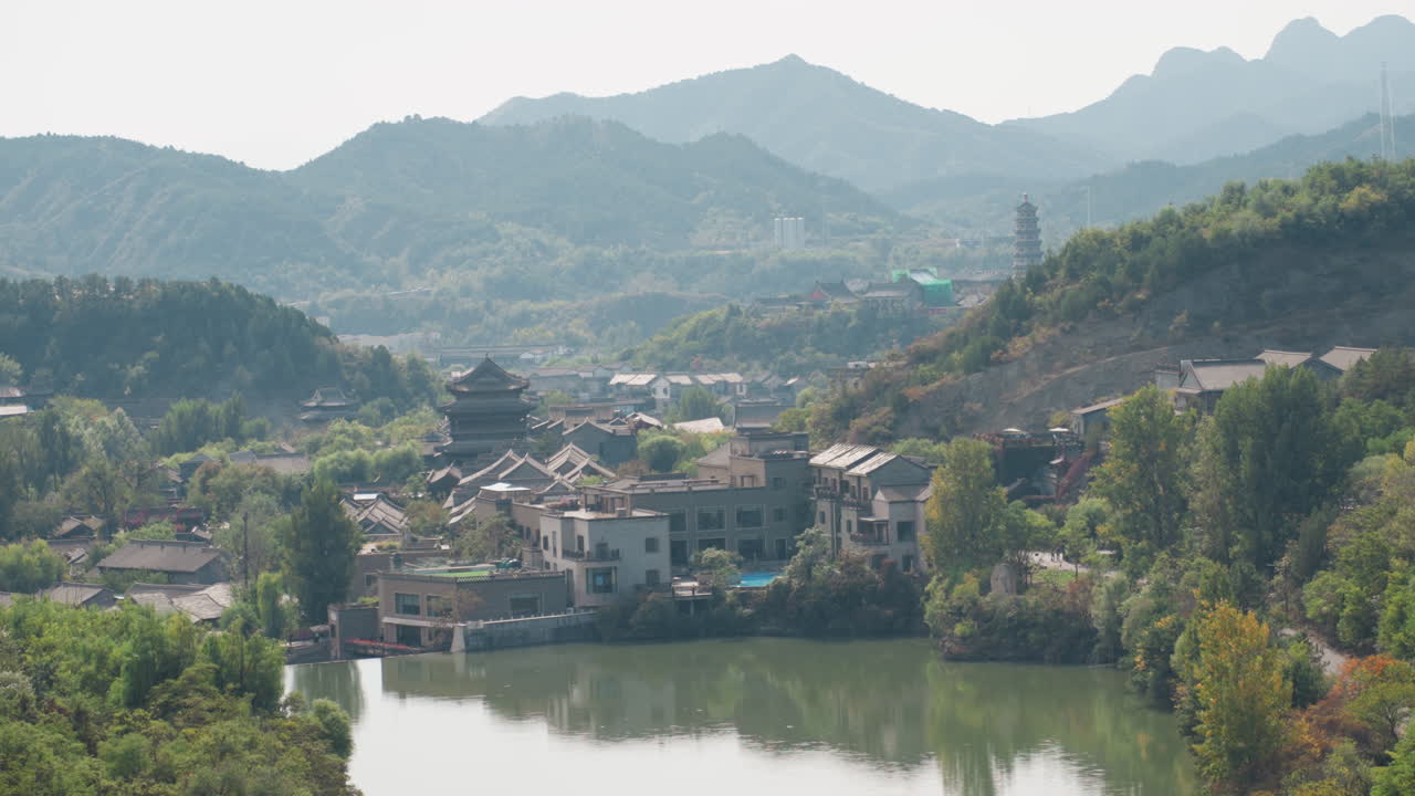 Villaggio panoramico sul fiume incorniciato dalle montagne. Veduta aerea di un insediamento tradizionale in collina con pagoda e tetti di tegole raggruppati, acqua calma e riflettente, alberi rigogliosi e pendii terrazzati sotto una luce soffusa