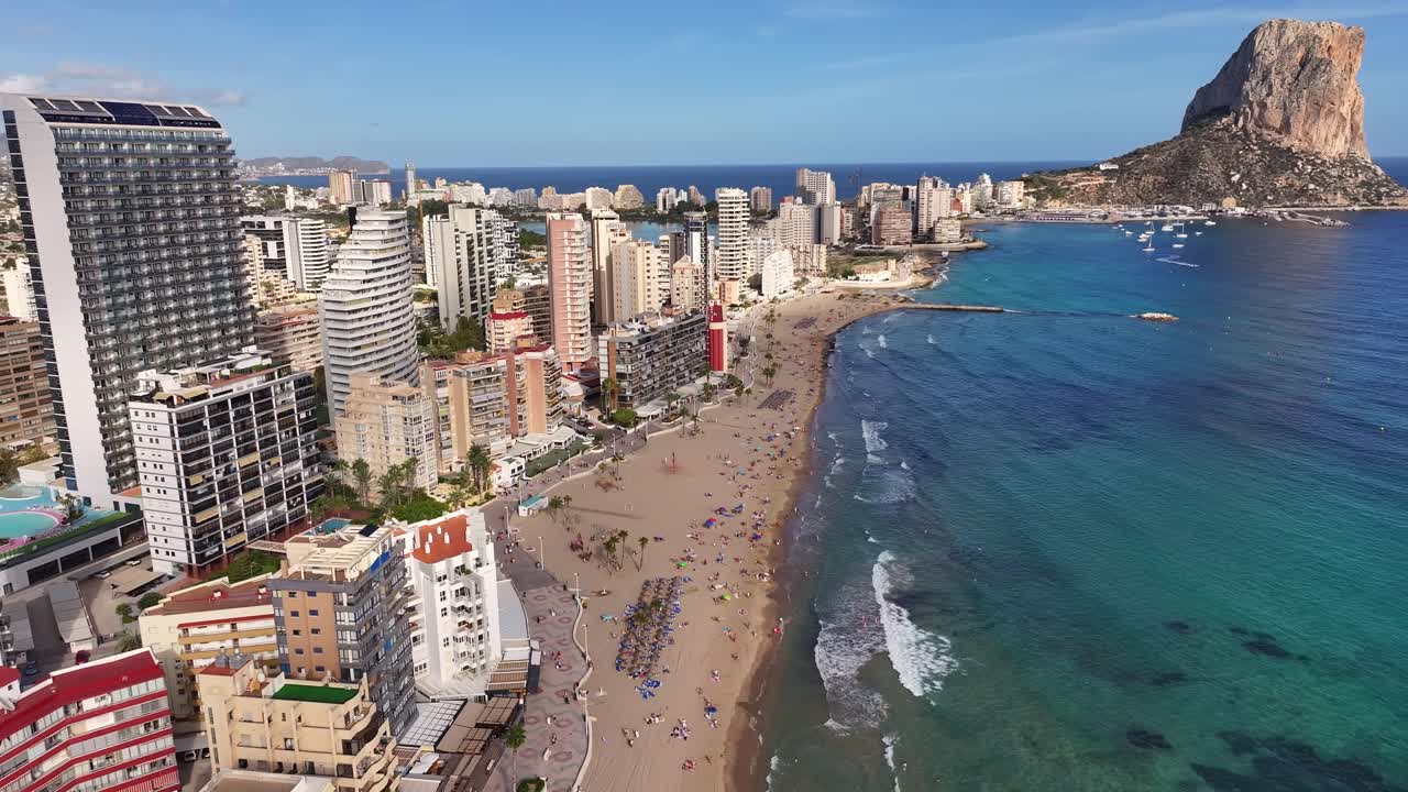 Aerial footage shows Calp’s sandy beach and skyline with high-rise buildings as the drone slowly pulls back, revealing the Peñón de Ifach and turquoise Mediterranean sea waters, sunny day