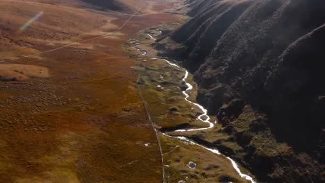 aerial drone footage flying over a small narrow water river stream at tibetan plateau during a sunny day with brown landscape on the left and tall mountains on the right. sun rays are visible on top.
