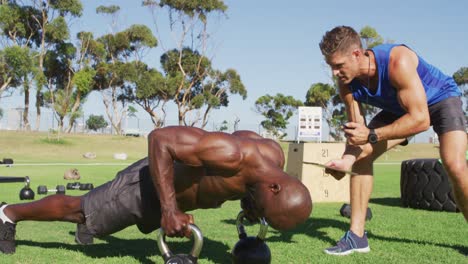 Two-diverse-fit-men-exercising-outdoors,-one-doing-push-ups-on-kettlebells-while-the-other-times-him