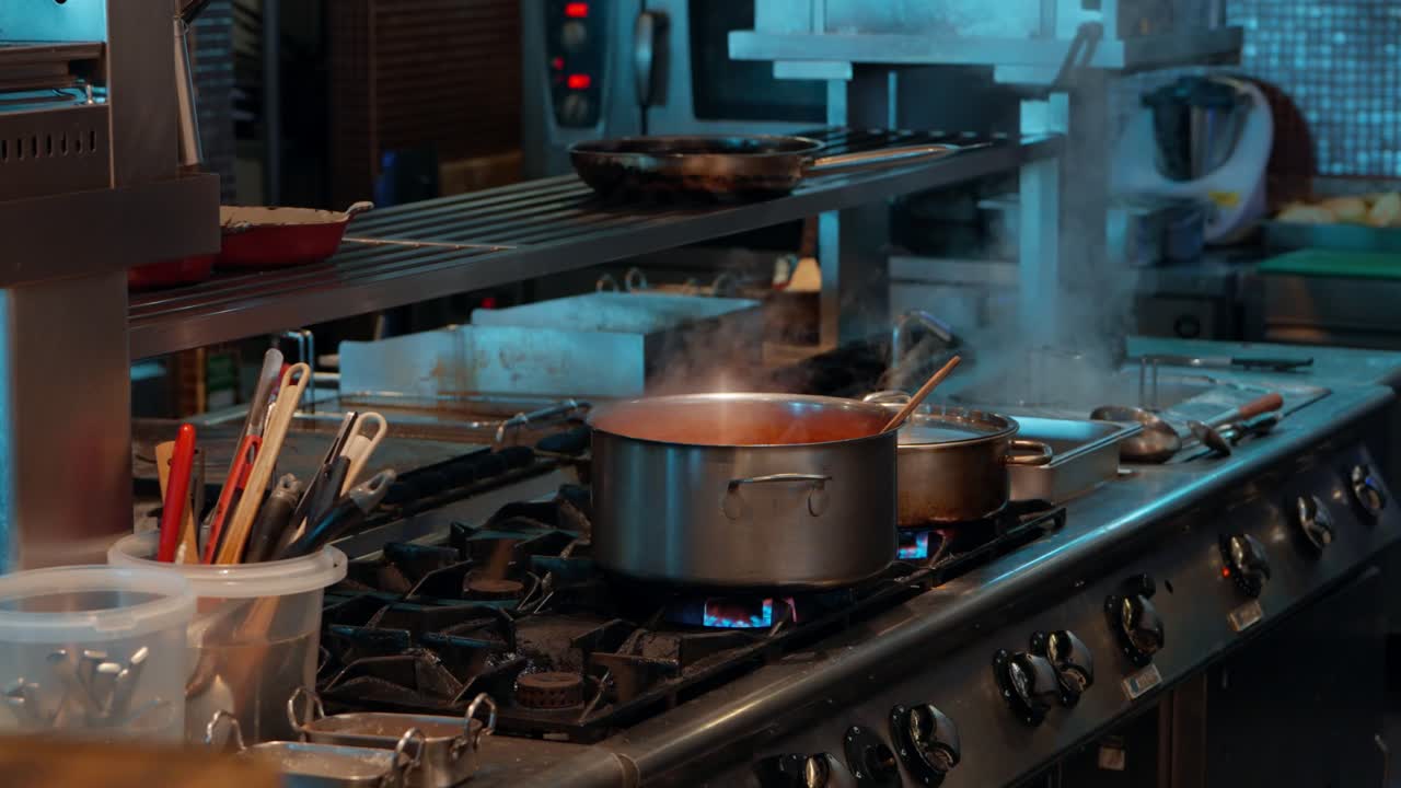 A wide shot of a busy professional restaurant kitchen showing a large pot of sauce simmering on a stainless steel gas stove while a chef works in the background, a behind-the-scenes culinary view