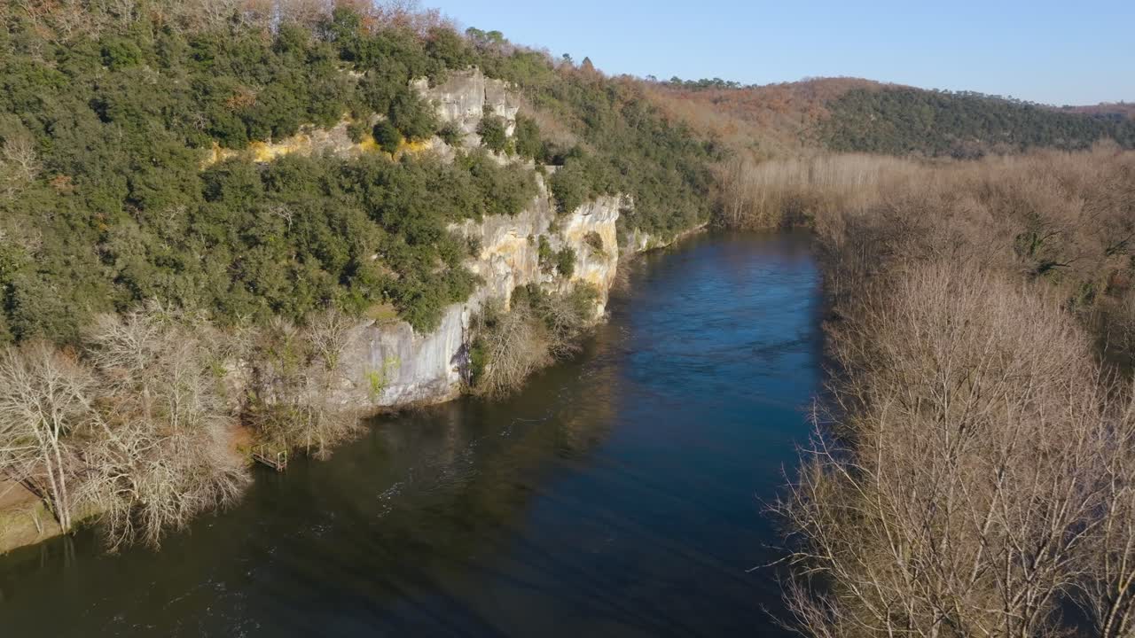 Záběr dronem z výšky na řeku Dordogne, která protéká kolem strmých vápencových útesů a holých stromů. Modrá voda se klidně vine francouzskou údolní krajinou v zimě.