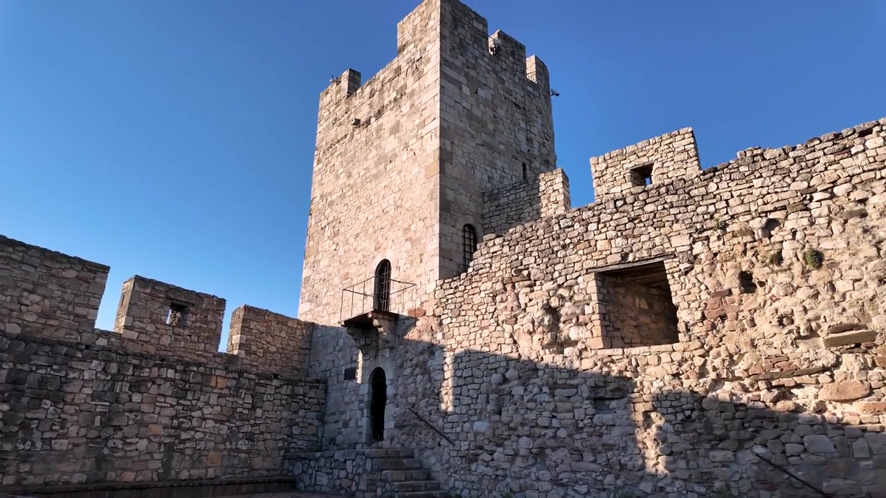 Il video mostra una visita a un antico castello di pietra. Gli spettatori possono vedere le robuste mura, le aree erbose e una torre sotto un cielo sereno. I gradini e le aperture del castello sono visibili in dettaglio