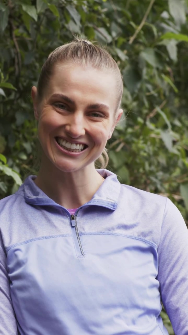 Vertical video: Smiling woman shifting into laughing in park with camera rolling in purple pullover