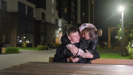 man seated using phone drops it to give attention to his lover who approaches from behind and gently wraps arms around his neck in romantic gesture amidst peaceful urban residential night backdrop