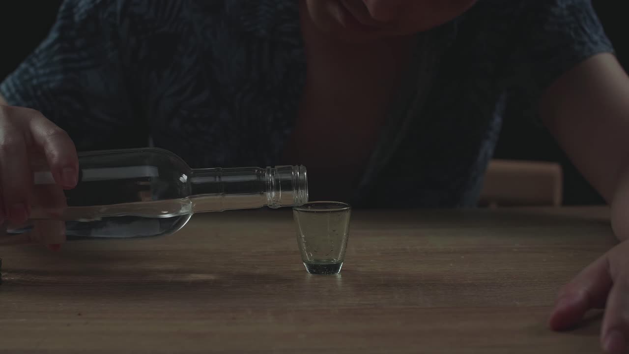 술에 취한 아시아인 남자가 술을 마시기 전에 글라스에 보드카를 부어 검은 바탕에서 잠을 자고 있습니다.