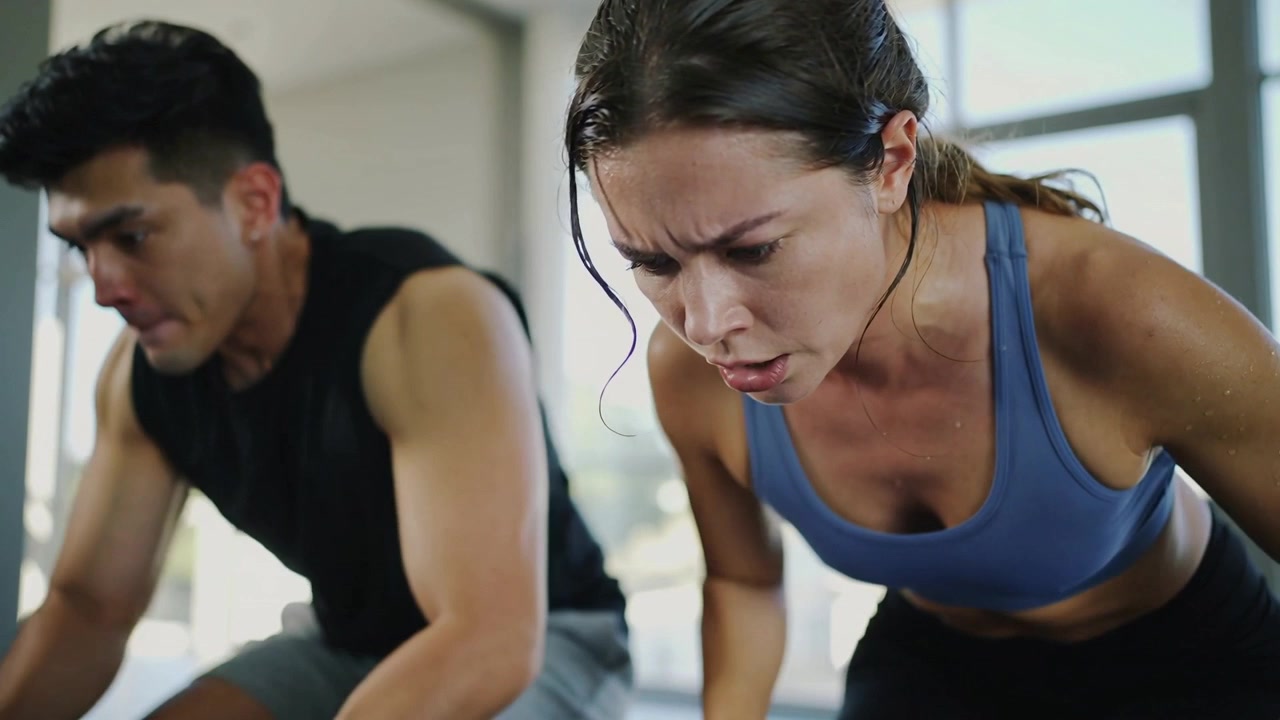Dos adultos pedaleando en bicicletas estáticas en un gimnasio, vistiendo ropa deportiva, con un reloj mientras entrenan.