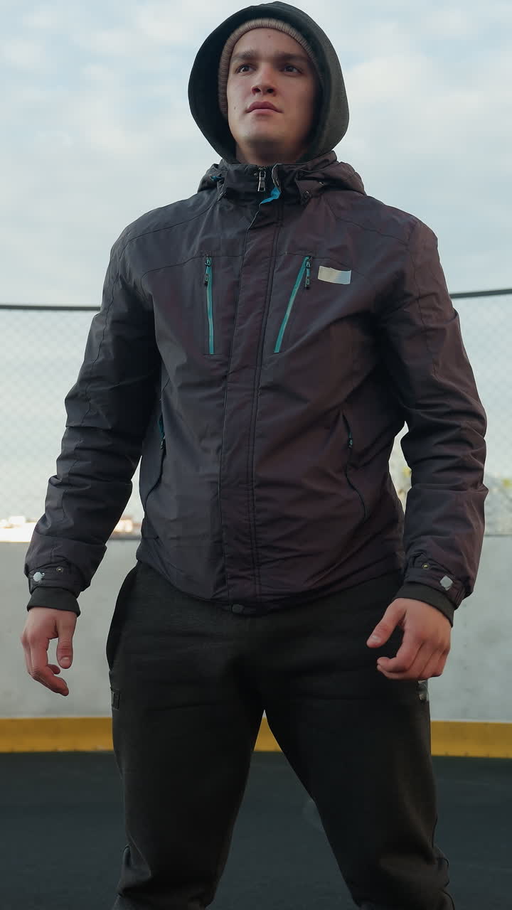 desportista realizando agachamentos profundos com as mãos fechadas, concentrando-se na preparação para o exercício em um campo de desporto ao ar livre, bola de futebol em primeiro plano, cercado por edifícios altos urbanos e poste de gol