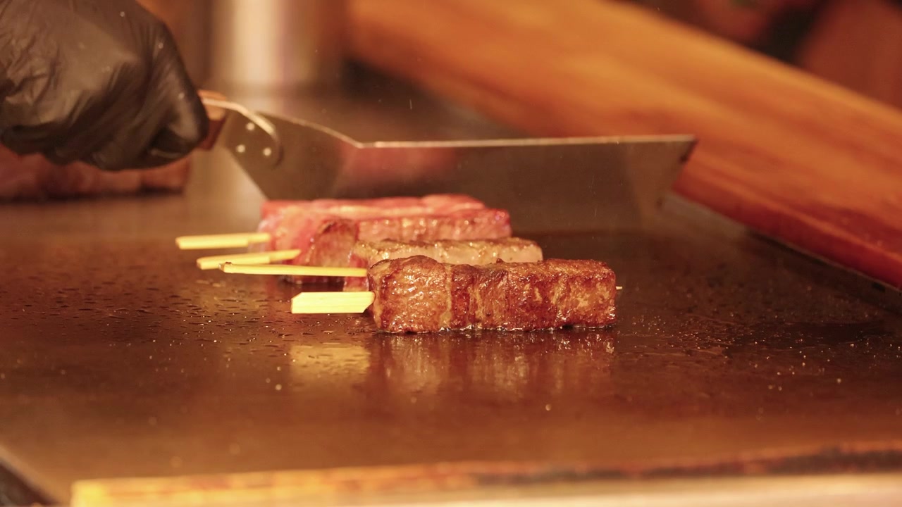 Primo piano di uno chef che usa una spatola per grigliare spiedini di manzo wagyu giapponese su una superficie teppanyaki calda