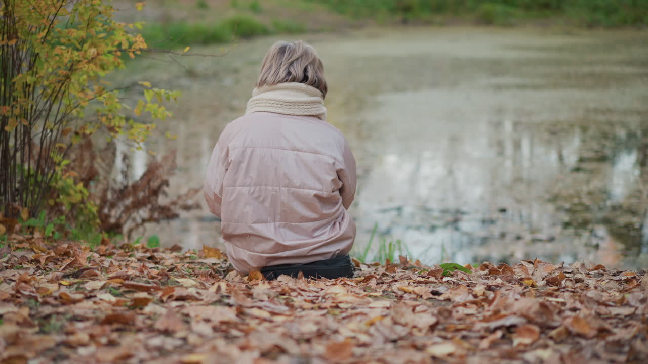 back view of woman wearing scarf and pink coat seated on ground near tranquil lake, surrounded by fallen autumn leaves, gazing at still water in quiet outdoor setting during serene seasonal moment