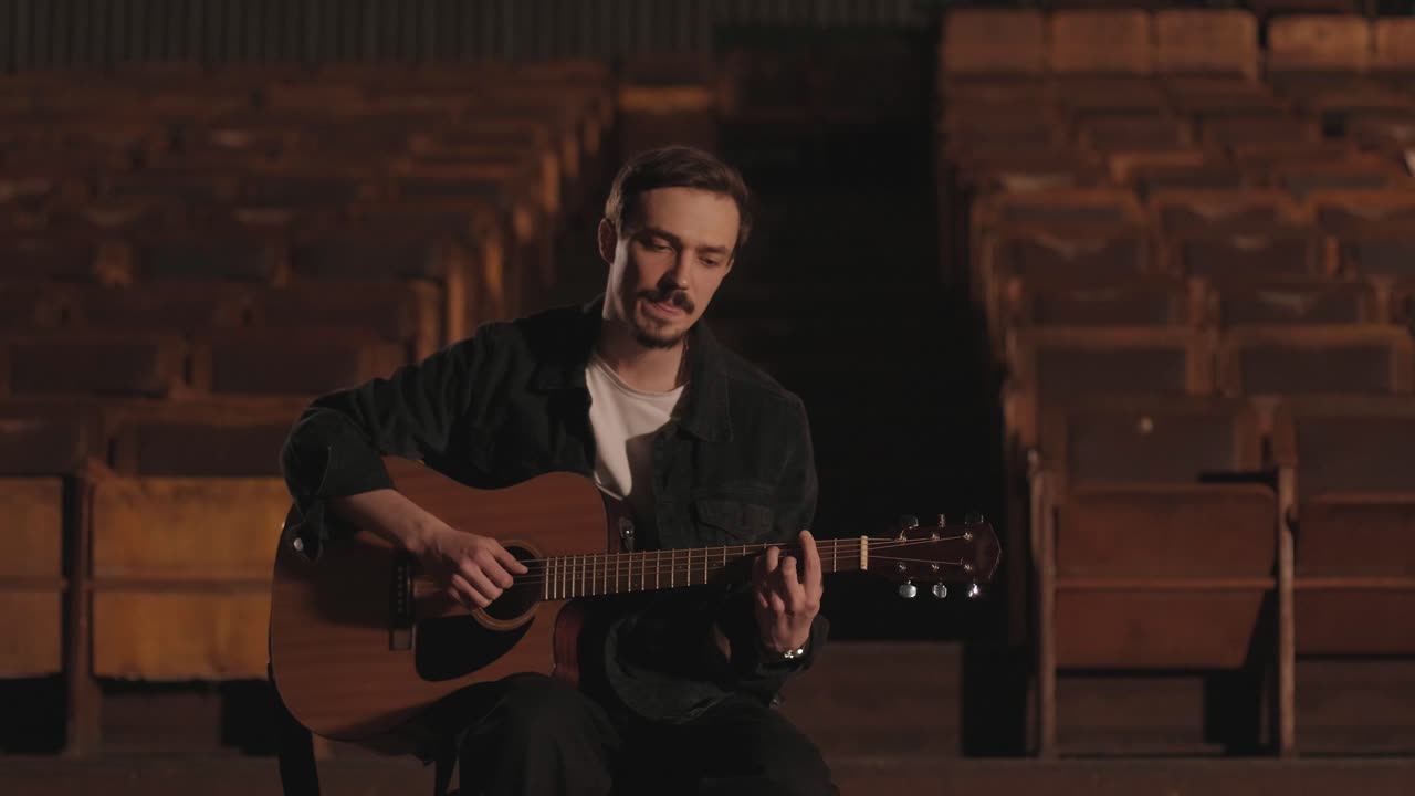 un chico guapo toca una guitarra acústica en un cine abandonado. el músico canta una canción y acompaña en la guitarra