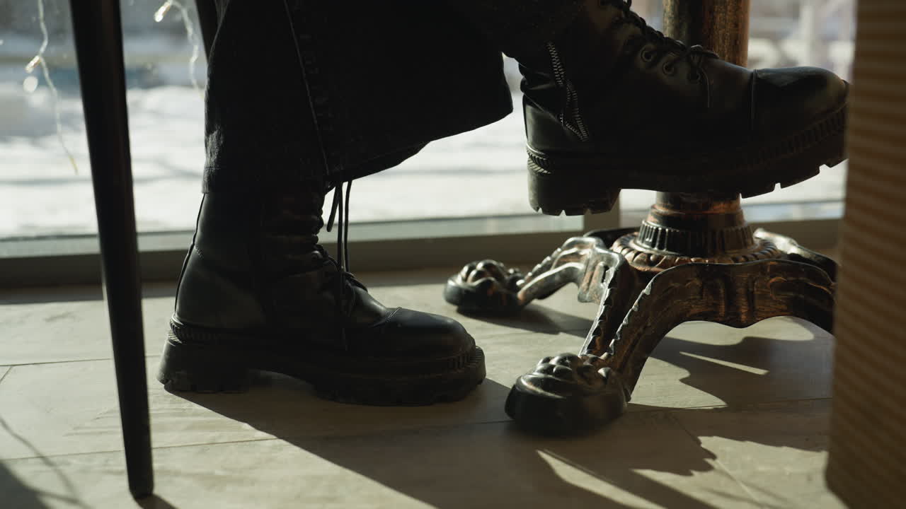 Close up leg view of lady seated indoors wearing black boots with laces beside decorative table leg, with sunlight casting shadows on tiled floor and snowy outdoor background seen through glass panel