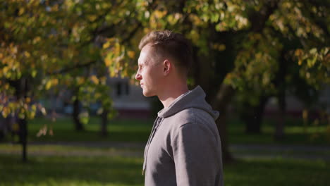 man in light gray hoodie captured in profile, with sunlight illuminating his face, standing against softly blurred autumn park background. neutral expression calmness in natural outdoor setting