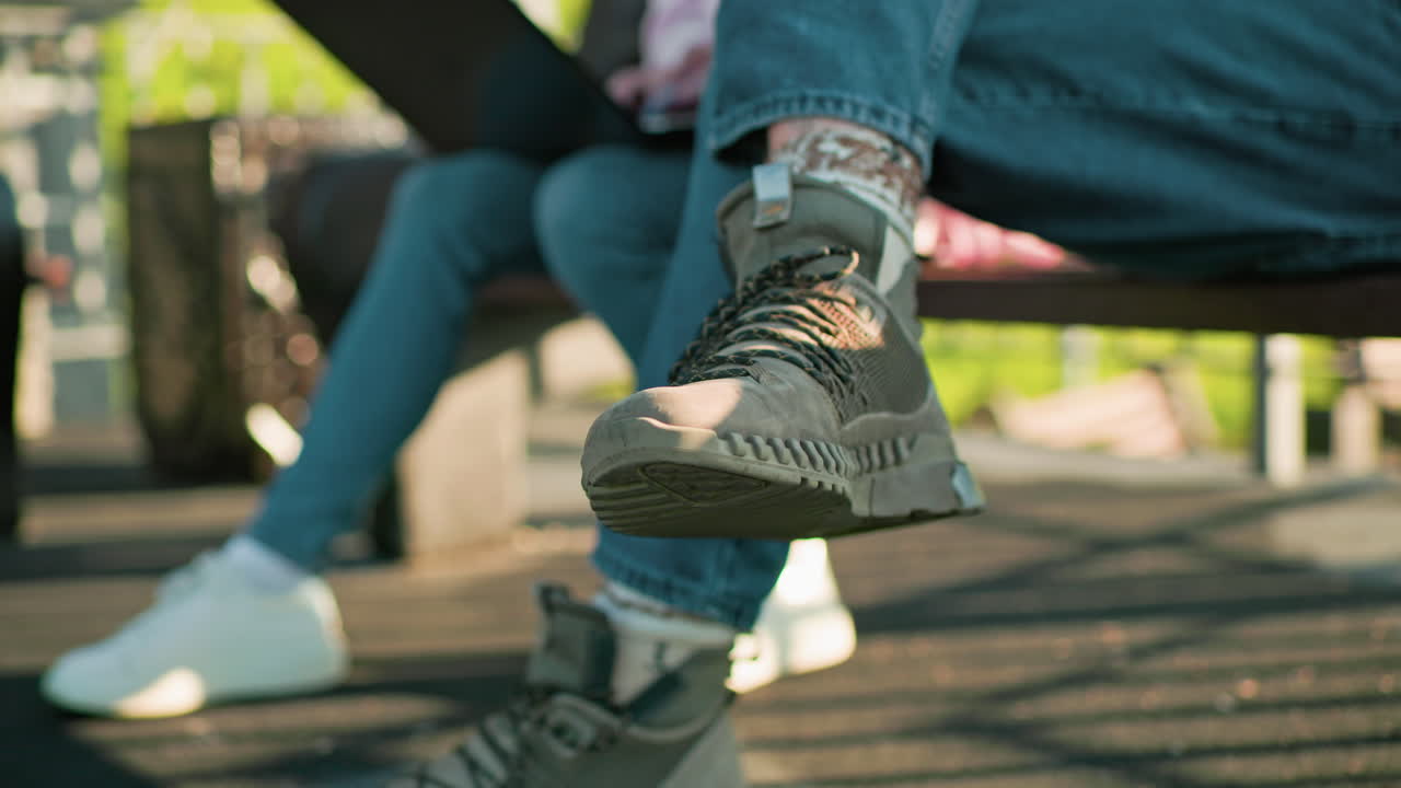 primer plano de las piernas de un hombre y una mujer sentados al aire libre en bancos de madera, con zapatillas deportivas y vaqueros casuales, uno usando una computadora portátil mientras la luz del sol proyecta sombras a través del entorno al aire libre relajado