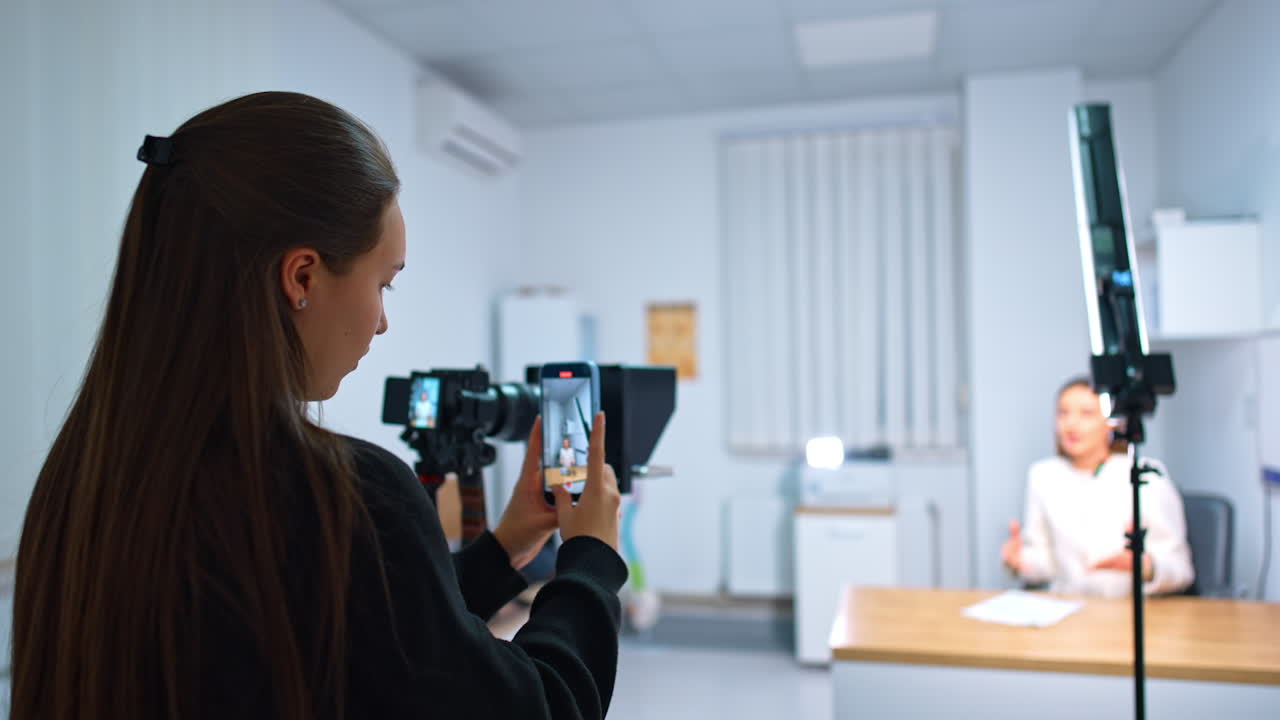 Rear view of a long-haired brunette girl taking video of a female speaker on the phone camera. Blogging concept. Blurred backdrop.